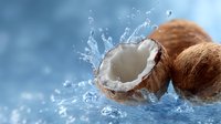 Fresh coconut halves with splashing water drops against blue background, dynamic food photography showing tropical fruit with white flesh and brown shell.
