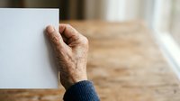 Senior male hand holding blank white card or paper against wooden floor background with natural window light, suitable for greeting cards, announcements, and promotional messaging.