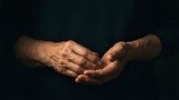Elderly hands cupped together in prayer or meditation pose against dark background, showing wrinkled skin and life experience in warm lighting.