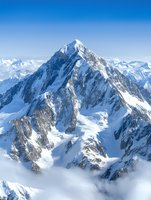 Majestic snow-covered mountain peak rising above clouds with dramatic rocky cliffs and glaciers against clear blue sky, capturing raw wilderness and alpine grandeur.