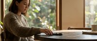 Woman in beige cardigan working at wooden desk near window with natural light, focused on laptop, suitable for business, productivity, and remote work concepts.