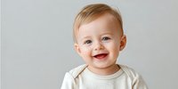 Happy Caucasian baby with light brown hair smiling against neutral gray background, wearing cream-colored onesie.