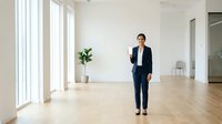 Young professional woman in business casual attire walking through modern, minimalist office space with natural light, wooden flooring, and potted plant for corporate environments.