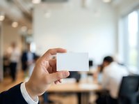 Businessman holding blank white business card in modern office environment with colleagues working in background.