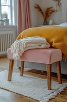Cozy pink upholstered bench with wooden legs and soft white throw blanket in bright modern bedroom interior with yellow bedding and pampas grass decor.