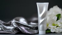 White cosmetic tube on dark background with silver silk fabric and white flowers, elegant composition for skincare and beauty product presentation.