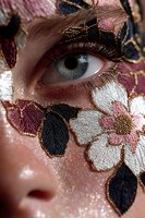 Close-up portrait of woman with intricate floral embroidery face art featuring metallic threads and beadwork around striking blue eye for fashion beauty concepts.
