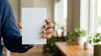 Unrecognizable person in casual blue shirt holding blank white card mockup indoors with blurred green plants and windows in background.