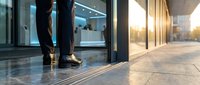 Businessperson in formal attire standing at modern glass entrance door of corporate office building during golden hour, entering workplace.