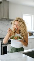 Happy blonde woman eating healthy quinoa salad in modern white kitchen, enjoying nutritious meal with fresh vegetables and greens for wellness lifestyle.