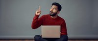 Young Hispanic man in red sweater pointing up with finger while using laptop computer, sitting cross-legged against gray wall with excited expression.