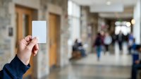 Close-up of hand holding blank white card in busy public corridor with blurred people walking in background, suitable for advertising, marketing mockups, and promotional campaigns.