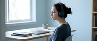 Young East Asian woman wearing headphones at desk with laptop and notebook, focused on online learning or remote work in bright modern office space.