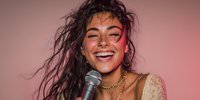 Young woman with messy hair laughing joyfully while holding microphone against pink background, wearing gold jewelry and light-colored top.