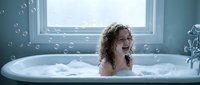 Happy young girl with curly hair laughing joyfully while taking bubble bath in white bathtub by window. Childhood hygiene and self-care routine concept.