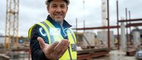 Male construction worker wearing safety helmet and high-visibility vest gestures welcomingly at active building site with cranes and steel framework in background.