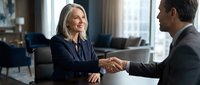 Professional mature businesswoman shaking hands with male colleague in modern office setting during successful meeting or partnership agreement.