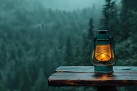Glowing vintage oil lamp on wooden surface against misty pine forest background in moody teal tones, creating atmospheric outdoor evening ambiance.