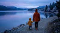 Father and son walking hand in hand along peaceful mountain lake shore during twilight hour with snow-capped peaks and evergreen forest reflecting in calm water.