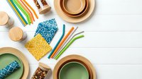 Flat lay arrangement of wooden bowls, colorful striped straws, patterned fabric napkins, and natural craft supplies on white surface for eco-friendly lifestyle content.
