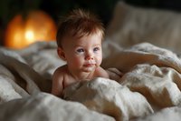 Adorable baby lying on white linen bedding in warm light, looking directly at camera with bright blue eyes and curious expression. Natural lifestyle portrait.