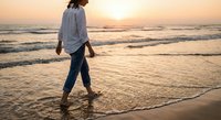 Young woman walking barefoot on sandy beach at golden sunset with ocean waves, peaceful solitude moment for wellness and relaxation concepts.