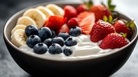 Fresh yogurt bowl topped with sliced bananas, ripe strawberries and blueberries in dark ceramic bowl, healthy breakfast macro photography.