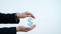 Young adult woman with fair complexion wearing a black blazer and decorative bracelet, holding a translucent dollar sign symbol between her hands against white background for financial concepts.