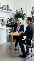 Senior businesswoman with gray hair presenting to male colleague in modern office meeting room with plants and artwork on shelves.