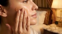 Young woman applying facial cream in cozy bedroom setting with warm lighting. Skincare routine, beauty treatment, moisturizing concept.