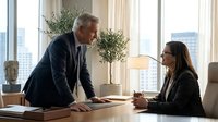 Mature male executive with gray hair and Caucasian ethnicity in dark business suit leaning over wooden desk during professional meeting with young female colleague of mixed ethnicity wearing glasses and dark blazer in modern high-rise office with city skyline view.