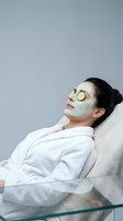 Young woman relaxing with green facial mask and cucumber slices on eyes in white bathrobe at spa for skincare and wellness treatment.