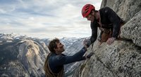 Rock climber helping teammate reach summit on mountain cliff with safety gear and ropes during outdoor adventure expedition in alpine wilderness landscape.