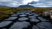 Dramatic stone pathway through volcanic landscape with distant erupting volcano, moody cloudy sky, and rugged terrain in remote wilderness area.