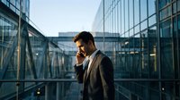 Professional businessman talking on phone in modern glass office building corridor during golden hour sunset lighting for corporate communications.