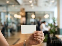 Unrecognizable person holding blank white card in modern office lobby with blurred colleagues and glass partitions in background.