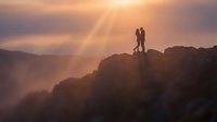 Couple silhouettes standing on mountain peak during golden sunset with dramatic light rays breaking through clouds creating romantic outdoor adventure scene.