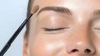 Professional makeup brush applying eyeshadow on closed eyelid of young caucasian woman with natural brows and clean skin during beauty procedure.