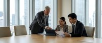 Senior male executive with gray hair and light skin tone, wearing dark suit, leads business meeting with two younger colleagues in modern high-rise office with city skyline view through floor-to-ceiling windows.