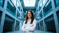 Confident young Caucasian businesswoman standing with arms crossed in modern corporate office building atrium, smiling professional portrait.