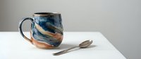 Handcrafted ceramic mug with swirled blue and brown glazed finish sits on white surface with wooden spoon, showcasing artisan pottery craftsmanship.