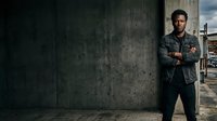 Young adult male with dark hair and facial stubble, wearing a dark denim jacket over casual clothing, standing confidently with arms crossed against a weathered concrete wall. Urban portrait for lifestyle, fashion, and professional use.