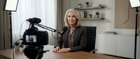 Middle-aged woman with blonde hair, Caucasian ethnicity, wearing professional brown blazer, seated at desk during video recording session with professional camera equipment and studio lighting setup.