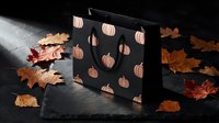Black shopping bag with copper pumpkin pattern surrounded by autumn maple leaves on dark surface for seasonal retail and fall marketing campaigns.