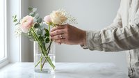 Woman arranging fresh pink roses and white flowers in glass vase on marble table by window. Floral arrangement, home decor, interior design concept.