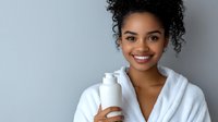 Young African American woman in white bathrobe holding skincare product bottle, smiling at camera against gray background for beauty and wellness concepts.