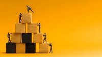 Small figurines of people working together to build a pyramid structure from wooden blocks against vibrant yellow background, symbolizing teamwork and achievement.