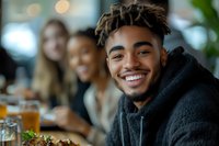 Young African American man with dreadlocks and facial hair smiling brightly at casual restaurant gathering with friends, enjoying food and drinks in warm ambient setting.