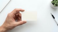 Hand holding blank white business card mockup on clean desk with pen and plant for branding presentation.