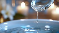 Crystal clear water drop falling from glass into rippling blue surface with soft bokeh lights and reflections in background, creating elegant splash effect.